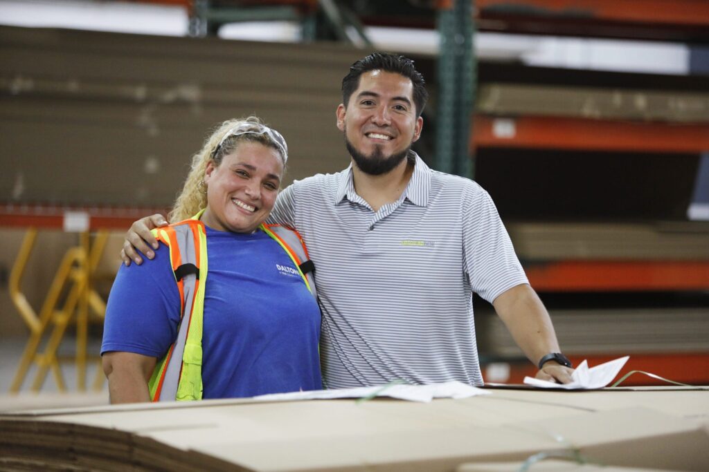 Smiling employees in a warehouse setting, wearing safety gear, representing teamwork in retail display and packaging solutions at The Royal Group.