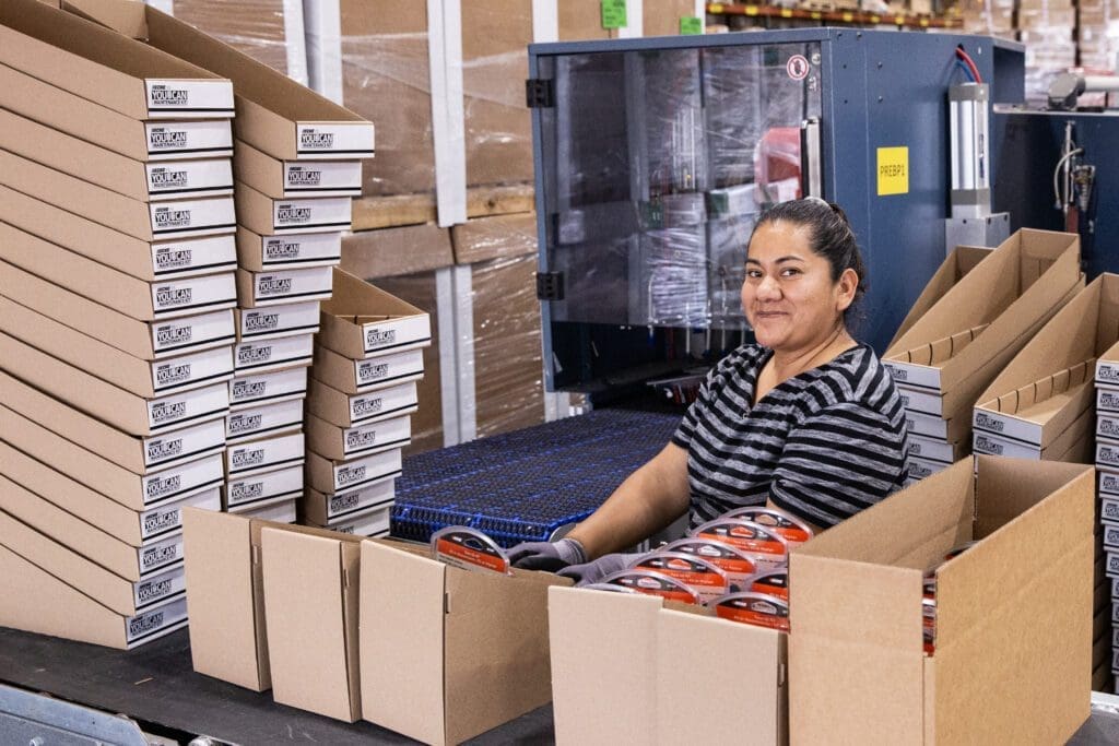 Worker smiling in fulfillment facility, surrounded by neatly stacked packaging boxes and ready-to-assemble containers, illustrating TRG's co-packing and fulfillment services.
